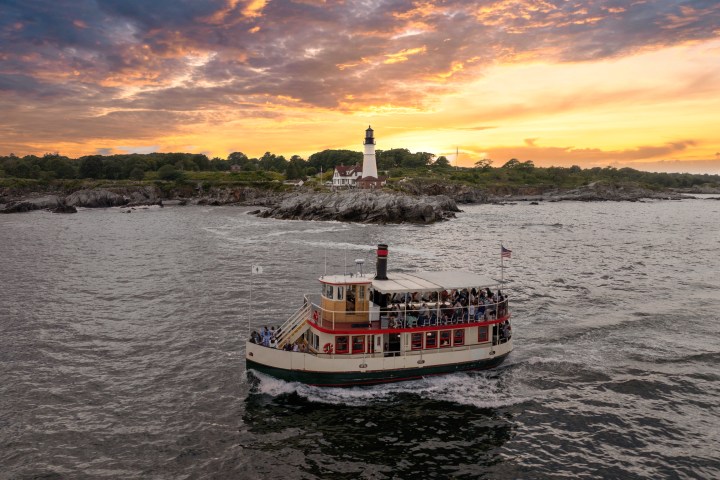 Boat in ocean near lighthouse at sunset with colorful sky.