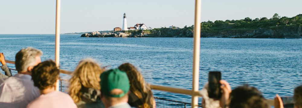 People on a boat photographing a distant lighthouse on a rocky shore.