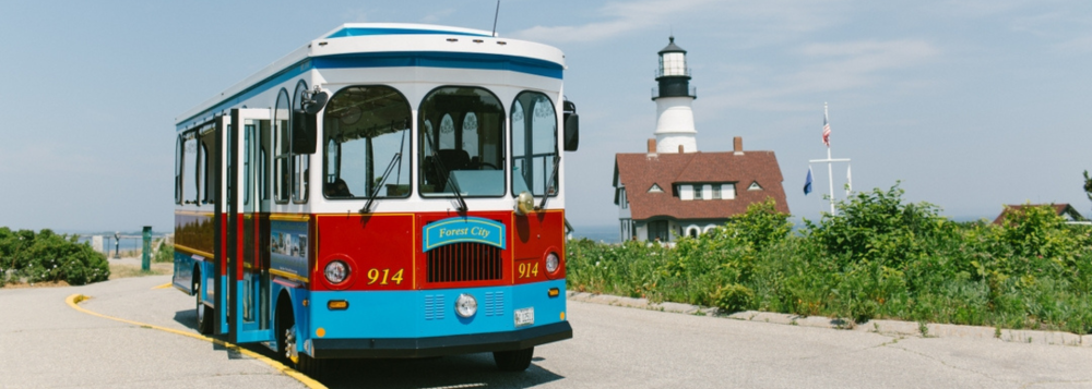 Colorful trolley bus near a lighthouse with a scenic coastal backdrop.