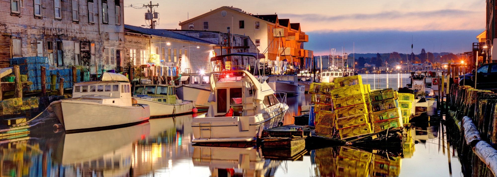 Rustic harbor at sunset with boats and stacked lobster traps along the dock.