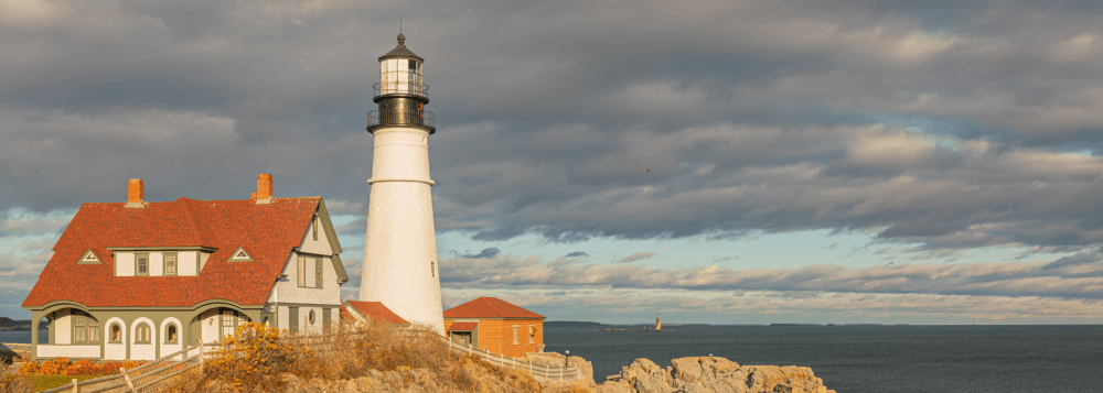 Lighthouse on rocky coast with house, cloudy sky, and ocean view.