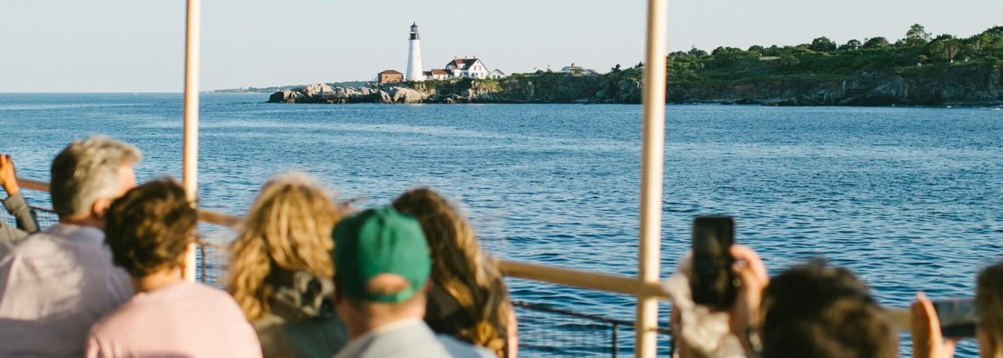 People on a boat looking at a distant lighthouse on a rocky shore across the water.