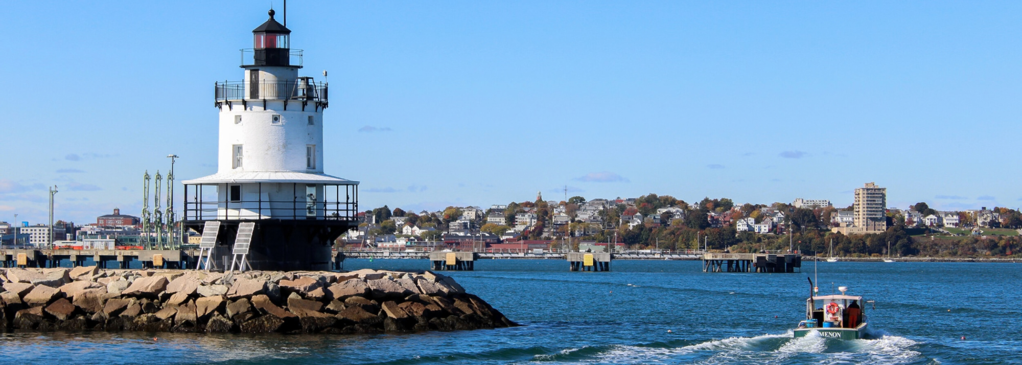 White lighthouse on rocky shore with a small boat in the water, town and blue sky in the background.
