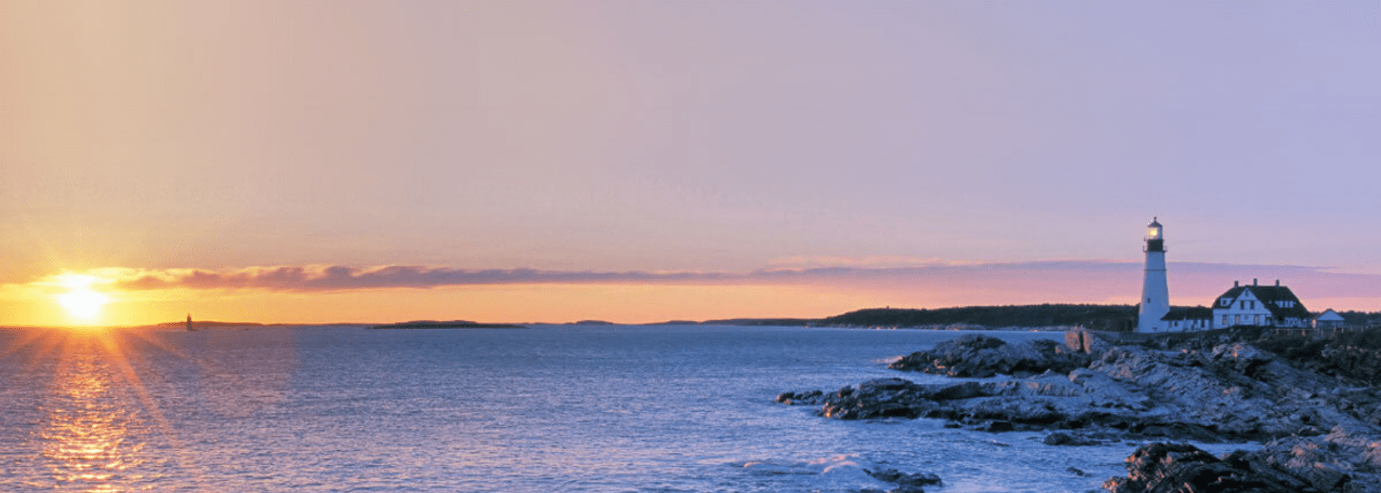 Sunset over ocean with a lighthouse on rocky shore.