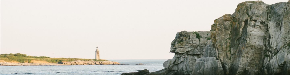 Distant lighthouse by the sea with rocky cliff in the foreground.
