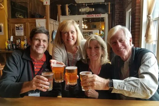 Four people smiling, holding glasses of beer in a cozy bar setting.