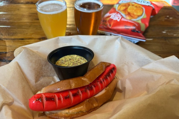 Red sausage in a bun with mustard, beer glasses, and spicy chip bag.