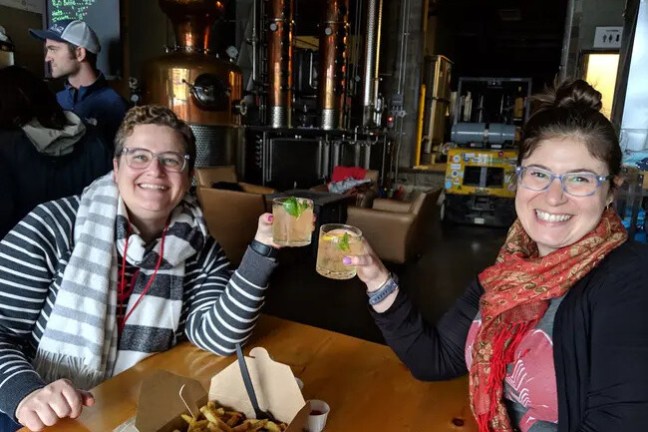 Two people smiling, toasting with drinks at a table with fries in an industrial setting.
